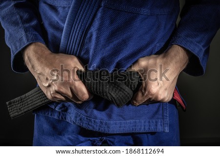 Close up on hand of unknown caucasian man holding brazilian jiu jitsu bjj black belt around his waist while wearing kimono gi in dark - martial arts mastery skill and confidence achievement concept