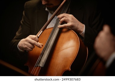 Close up on hand of unrecognizable devoted male musician strumming cello while playing with chamber quartet in dramatic light, focus on fingers and strings