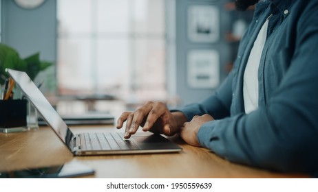 Close Up On Hands Of A Black African American Man Working On Laptop Computer While Sitting Behind Desk In Cozy Living Room. Freelancer Working From Home. Browsing Internet, Using Social Network.