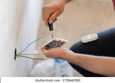 Close up on hands of caucasian man electrician holding screwdriver working on the plug electric on residential electric system installing white AC power socket on gray wall at home repair close up