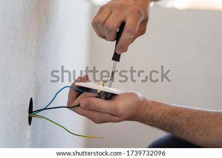Close up on hands of caucasian man electrician holding screwdriver working on the plug electric on residential electric system installing white AC power socket on gray wall at home repair close up