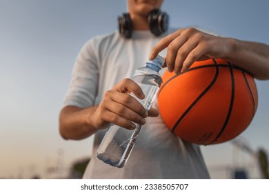 close up on hands midsection of unknown caucasian man teenager open plastic bottle of water outdoor in sunny day doing sports with basketball