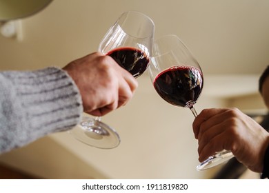 Close Up On Hands Of Two Unknown Caucasian Men Holding Glasses With Red Wine Toasting - Man Cheers Celebrating While Holding Drink At Home Or Restaurant