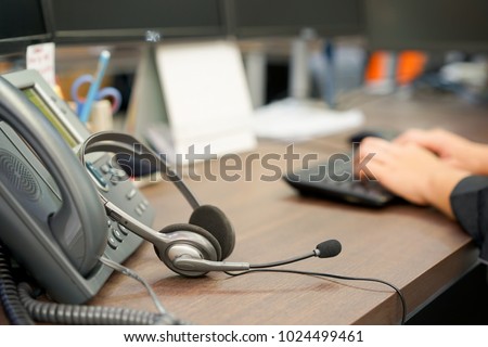 close up on headset of call center leaning on telephone with blur network engineering typing on keyboard for reply case to customer or monitoring alarm , technology concept
