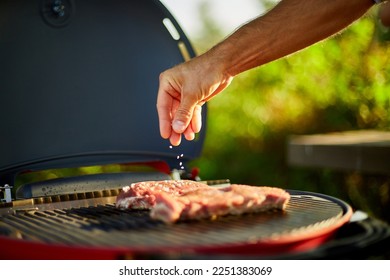 Close up on man's hand seasoning meat on the gas grill on barbecue grill outdoor in the backyard, grilled roasted steak meat, summer family picnic, food on the nature.