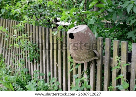 A close up on an old, rusty and used up jug or mug ganging from a rural wooden fence located next to a highly overgrown orchard or garden seen on a sunny summer day in Poland