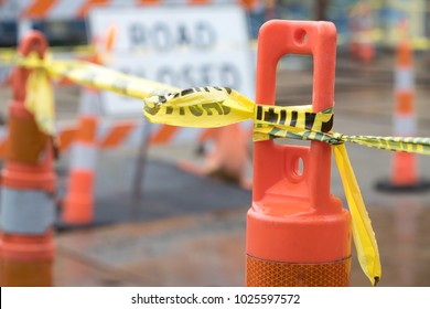 Close up on an orange construction cone with yellow caution tape, at a road construction site, with a Road Closed sign and barrier in the blurry background - Shutterstock ID 1025597572
