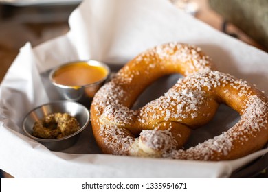 Close Up On A Salted Soft Pretzel In A Parchment Paper Lined Basket, With Mustard And Cheese Dipping Sauces