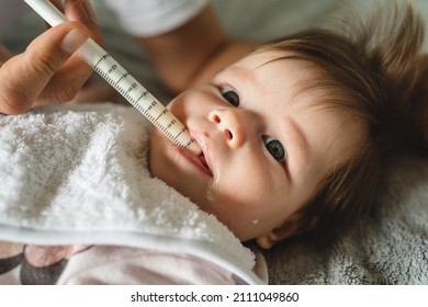 Close Up On Small Caucasian Baby Four Months Old And Hand Of Unknown Father Giving Her Medicine Antibiotics While Lying On Bed At Home Parenthood And Health Issues Liquid Drugs Oral Syringe Concept