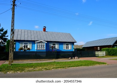 A close up on a small wooden abandoned house with slanted roof made out of metal and a wooden fence located next to an asphalt roaf and a power pylon seen on a sunny summer day in Poland
