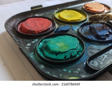 Close Up On A Water Paint Palette For Kids Made Of Plastic With Dry Cracked Paint And And Wooden Paint Brush On Top. Green, Yellow, Red, Blue, Orange Colors