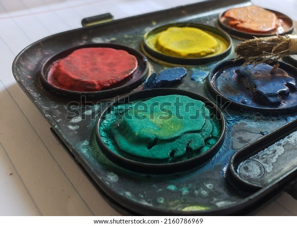 Close up on a water paint\
palette for kids made of plastic with dry cracked paint and and\
wooden paint brush on top. Green, yellow, red, blue, orange\
colors
