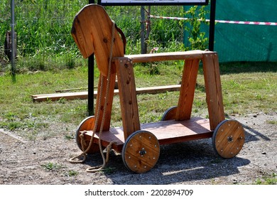 Close Up On A Wooden Toy Resembling A Horse With A Seat Made Out Of Plank And Some Wooden Wheels Attached To It Seen In The Middle Of A Public Park Standing On A Gravel Pavement On A Sunny Summer Day