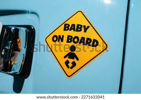 Close up on a yellow sticker in the shape of a warning sign with the inscription 'Baby on Board' stuck to the back of a turquoise colored car