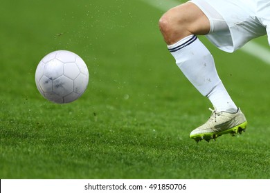 Close up one leg and feet of football player in white socks and light gray shoes running and dribbling with the ball playing on green grass pitch with dust water splashes