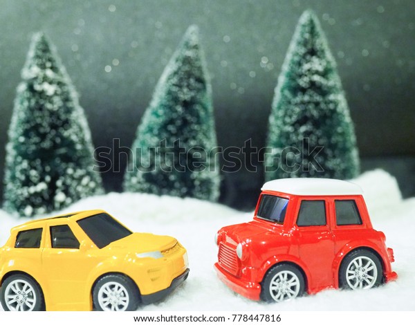 A close up of\
one red toy car and one yellow toy car on snow with blurred\
Christmas tree in the\
background.