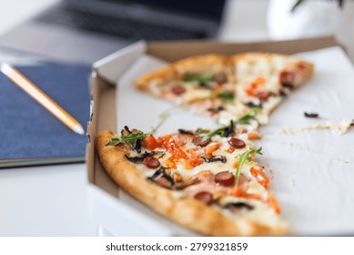 Close up open pizza box with a slice in focus on a desk near a notebook and laptop for food background and work lunch banner.