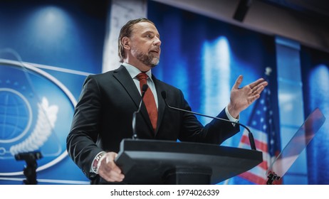 Close Up Of Organization Representative Speaking At A Press Conference In Government Building. Press Officer Delivering A Speech At A Summit. Minister At Congress. Backdrop With American Flags.