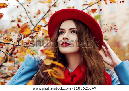 Close up outdoor portrait of young beautiful happy smiling girl wearing red hat and scarf posing near autumn tree. Model with red lips, long hair. Lady looking up.