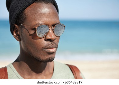 Close Up Outdoor Shot Of Handsome Bearded Young African American Male Model Wearing Black Hat And Mirror Lens Sunglasses, Posing Against Blurred Marine Landscape Background, Carrying Backpack