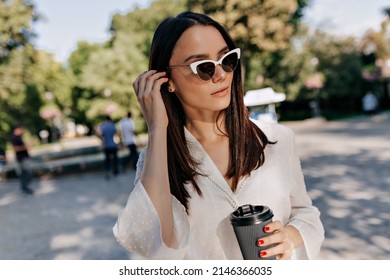 Close Up Outside Photo Of Pretty Woman In Sunglasses And Wearing Elegant Blouse Is Touching Hair And Looking Aside On Green Park Background. 