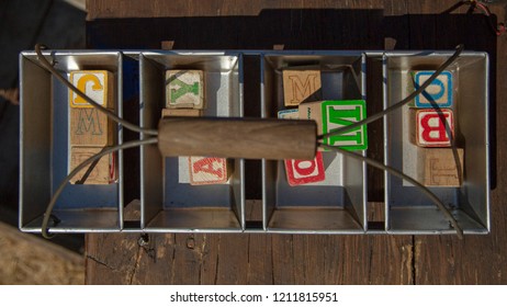 Close Up Overhead Perspective Of A Collection Of Old Children's Playing Blocks In A Metal Box With A Wooden Handle Against An Out Of Focus Wooden Table Background
