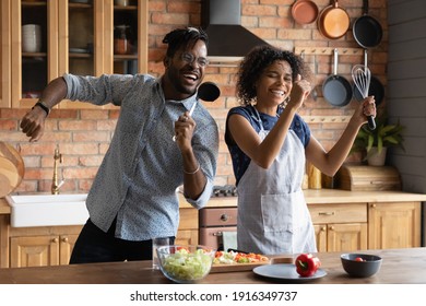 Close up overjoyed African American couple singing into kitchenware, having fun, dancing, listening to music in kitchen, cooking salad, excited wife and husband holding whisk and ladle a microphone