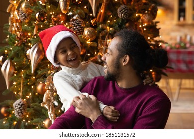 Close up overjoyed African American father and little son wearing festive red cap celebrating Christmas, laughing dad and child hugging, sitting near decorated tree at home, having fun together - Powered by Shutterstock