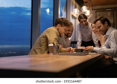 Close up overjoyed diverse colleagues looking at phone screen together, sitting at wooden table in office, having fun at late meeting, watching funny video, laughing at joke, making group video call