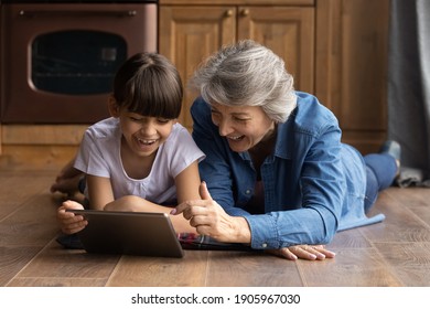 Close up overjoyed mature grandmother with little granddaughter using tablet, having fun, lying on warm wooden floor with underfloor heating at home, enjoying leisure time with gadget, browsing apps - Powered by Shutterstock