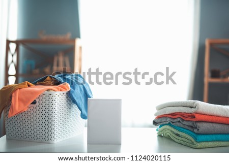 Close up of pack of detergent on white table. Wicker clothesbasket full of dirty linens and pile of folded towels are aside