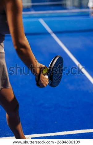 Close up of a padel player getting ready to serve the ball holding a black padel racket with a blue padel court in the background.