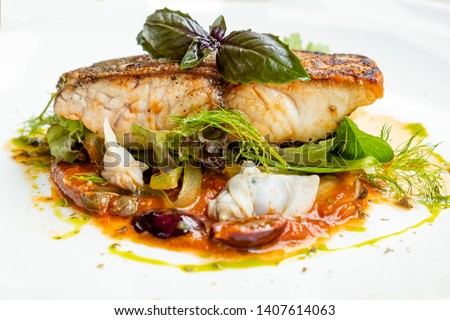 close up of a pan seared grill seabass fish fillet italian  livornese sauce with tomato, olives, capers, dill, oregano, chilly, clams , parsley, onion , basil, in a white plate , natural light 