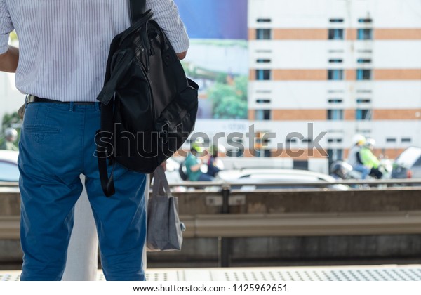 Close up a part of back a man\
wearing light shirt and navy pants blue carry black bag waiting for\
train in rush hour with cars full on the road\
background.