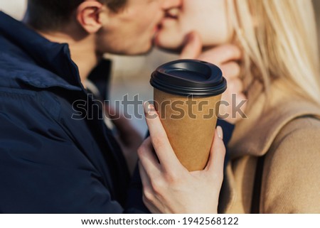Close up of passionate kissing man and woman while walking outdoor. They are drinking hot beverage.