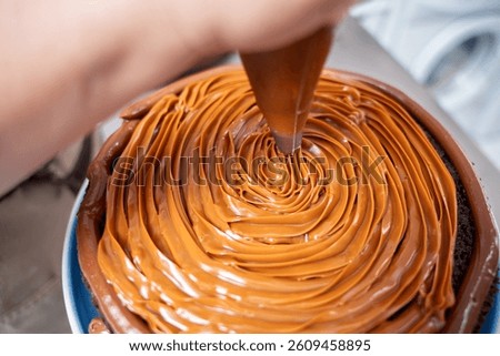 Close up of a pastry chef using a piping bag, creating a swirling design of dulce de leche frosting on a rich chocolate cake