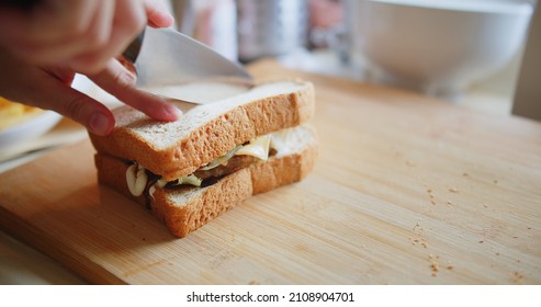 Close Up Person Cuts Sandwich Bread With Meat, Cheese And Vegetables In Half By Sharp Knife On Wooden Board At The Kitchen, Making Of Fast Food At Home	
