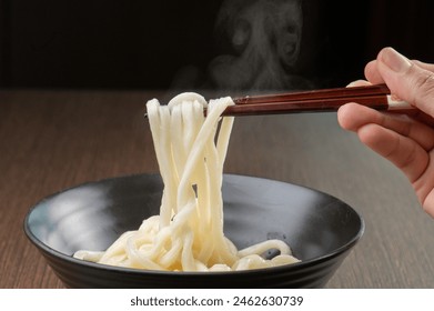 A close up of a person holding chopsticks with steaming udon noodles over a black bowl. - Powered by Shutterstock