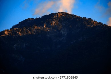 Close perspective of a formidable mountain summit bathed selectively in golden sunset light while the lower rocky slopes remain hidden deeply within dark dramatic shadows