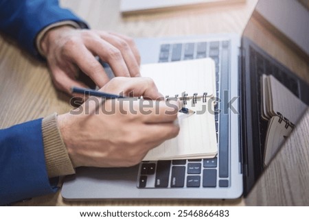 Close perspective of a man's hand making entries in a notepad on a cutting-edge laptop, with a soft blur in the surrounding