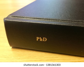 Close up of PhD thesis with black binding and letters PhD in gold leaf on the spine lying on a wooden surface. Landscape orientation.