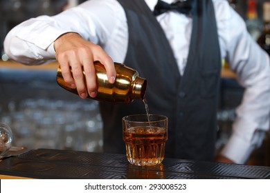 Close Up Photo Of A Bartender Holding A Golden Shaker In His Hand And Pouring A Cocktail In A Low Wide Glass, Shelves Full Of Bottles With Alcohol On The Background