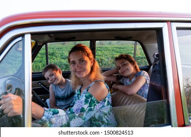 Close Up Photo Of A Beautiful Young Blonde Mom In Dress With Flowers Print Driving A Car With Her Teen Children, Enjoying Traveling Together, Summer Holidays And Vacation Concept