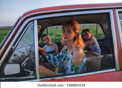 Close Up Photo Of A Beautiful Young Blonde Mom In Dress With Flowers Print Driving A Car With Her Teen Children, Enjoying Traveling Together, Summer Holidays And Vacation Concept