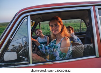 Close Up Photo Of A Beautiful Young Blonde Mom In Dress With Flowers Print Driving A Car With Her Teen Children, Enjoying Traveling Together, Summer Holidays And Vacation Concept