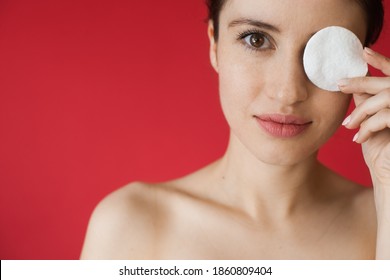 Close Up Photo Of A Caucasian Unclothed Woman With Freckles Posing On A Red Background While Covering Eye With A White Disk