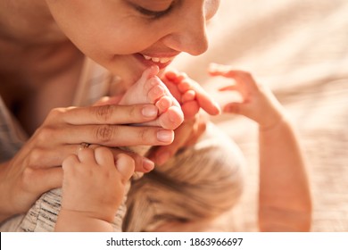 Close Up Photo Of The Cheerful Caucasian Woman Holding Legs Of Her Baby Near Her Face And Looking At Him With Tenderness While Spending Time At Bedroom