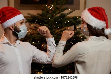 A Close Photo Of Elbow Greeting To Avoid The Spread Of Covid-19 Against The Background Of A Christmas Tree At Home. A Man And A Woman In Medical Masks And Santa Hats Instead Of A Hug, They Bump Elbows