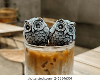 Close up photo of a glass of ice cold latte topped with two owl shaped chocolate cube at the surface in a cafe with sofa chair in the background - Powered by Shutterstock