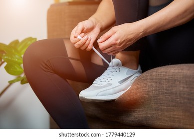 A Close Photo Of The Hands And Legs Of A Sporty Girl In Black Tights Who Is Tying Her Shoelaces On White Sneakers While Sitting On The Couch At Home.